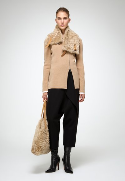 Donna che indossa un maglione beige con zip e collo in pelliccia oversize, pantaloni neri a taglio corto, stivaletti neri con tacco e porta una borsa tote in pelliccia beige.
