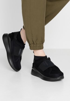 Person iført olivengrønne joggebukser og sorte slip-on sneakers med tykke såler og brede elastiske stropper, stående på hvidt gulv.