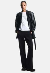 Giubbotto di pelle nera sopra una maglietta bianca con testo, abbinato a pantaloni larghi neri e scarpe nere. Colori solidi, design casual.
