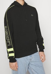 Svart hoodie med neongröna detaljer på ärmarna och logotypen. Har en dragsko i huvan och en avslappnad passform.