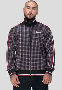 Marynarka sportowa w czarno-czerwono-białą kratę. Posiada wysoki kołnierz, zamek z przodu oraz paski na rękawach. Haftowane logo na piersi.