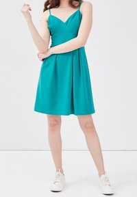 Robe A-line turquoise avec des bretelles spaghetti, décolleté en V et jupe plissée. Portée avec des baskets blanches, sur un fond clair.
