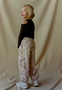 Juodas off-shoulder viršus, šviesios raštuotos cargo kelnės rožinės ir žalios spalvos, elastinga juosmens juosta, kišenės ir baltos sportbačiai. Modelis žiūri atgal.