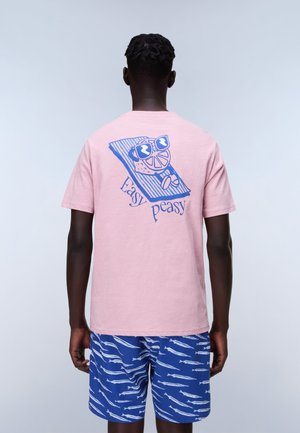 Homme portant un t-shirt rose avec le texte "Easy Peasy" et une illustration de tranche de citron sur une serviette rayée, associé à un short bleu à motifs de poissons.
