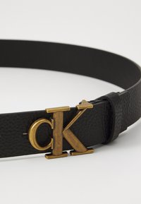 Fekete bőr öv, texturált felülettel és aranyszínű "ck" csattal. A dizájn a klasszikus és minimalista esztétikát hangsúlyozza.