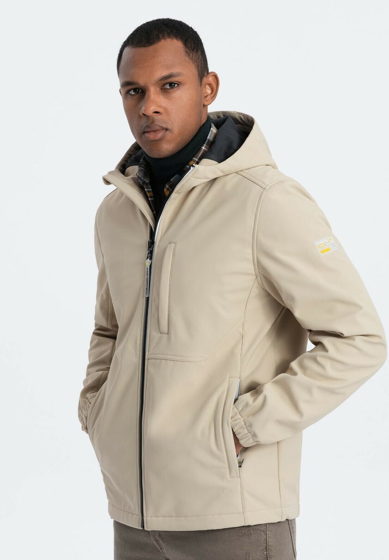 Beigefärgad väderbeständig jacka med huva, dragkedja framtill och elastiska ärmslut. Har en rutigt foder och sidofickor. Slät textur.