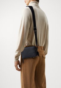 Liten, svart lær crossover-veske med teksturert overflate, med glidelås og justerbar stropp, båret over en beige genser.