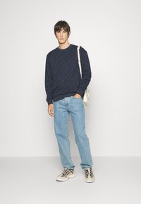 Felpa blu navy con motivo a diamante testurizzato, abbinata a jeans azzurri. Il modello indossa sneakers leggere e porta una borsa piccola.