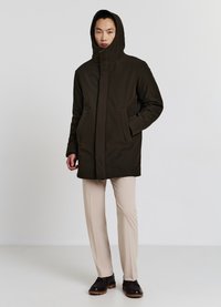 Cappotto verde scuro con cappuccio, chiusura frontale con zip e due tasche laterali; abbinato a pantaloni beige e scarpe marroni. Tessuto tinta unita.