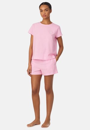 Mujer de pie, descalza, vistiendo una camiseta de manga corta a rayas en rosa claro y blanco, y pantalones cortos a juego, con las manos en los bolsillos, fondo neutro.