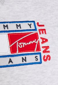 Parche con el logo de Tommy Jeans bordado en rojo, blanco y azul sobre un fondo de tela gris; presenta letras en negrita y una firma estilizada.