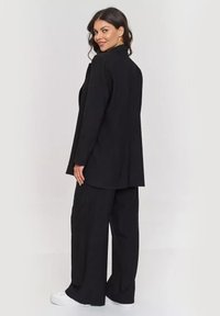Czarny dopasowany żakiet i spodnie w szerokiej nogawce z gładkiego materiału. Minimalistyczny design z prostymi liniami i otwartym tyłem. Białe buty.