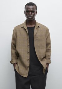 Braun karierter Button-Up-Hemd mit lockerer Passform, langen Ärmeln und einer Brusttasche, über einem schwarzen Rundhalsausschnitt-T-Shirt getragen.