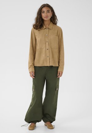 Femme aux cheveux bouclés portant une chemise marron à boutons, un pantalon cargo ample vert foncé et des chaussures plates marron, debout devant un fond uni.