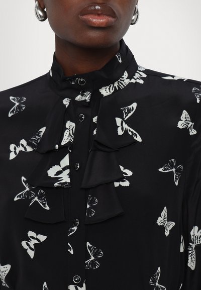 Blusa nera con motivo a farfalla bianca, colletto alto, dettagli frontali arricciati e chiusure a bottone. Tessuto liscio con una finitura lucida.