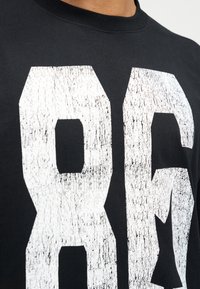 Μαύρο T-shirt με μεγάλα, φθαρμένα λευκά νούμερα "86" στο μπροστινό μέρος. Σχεδιασμός με στρογγυλή λαιμόκοψη και απαλό ύφασμα.