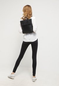 Zwarte leren rugzak met een omgevouwen klep, minimalistisch ontwerp, gladde textuur, gedragen met een wit shirt en zwarte broek.