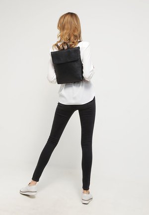 Black leather backpack with a fold-over flap, minimalistic design, smooth texture, worn with a white shirt and black pants.