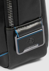 Zwarte leren tas met een hoek, voorzien van een zilveren logo plaat, blauwe accenten en een zichtbare rits. Textuur oppervlak en stiksel detail.