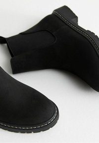 Bottes cheville en daim noir avec panneaux latéraux élastiques, bouts arrondis et couture blanche contrastante le long de la semelle. Semelle extérieure en caoutchouc robuste.