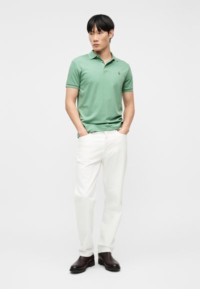 Άνδρας που φοράει γαλάζιο-πράσινο πόλο πουκάμισο, λευκό παντελόνι και σκούρα καφέ μπότες, στέκεται με τα χέρια στις τσέπες μπροστά σε λευκό φόντο.