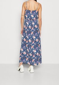 Robe maxi en tissu bleu foncé avec un motif floral rose et violet. Présente des bretelles fines, une taille froncée et une silhouette fluide. Chaussures à talons blocs blanches.