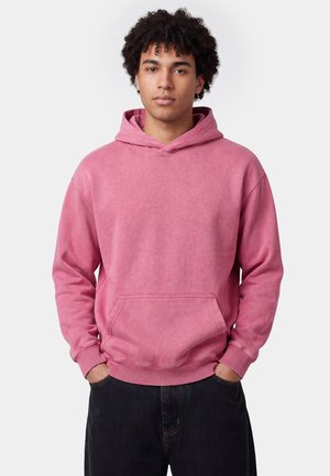Junger Mann mit lockigem Haar, der einen rosa Kapuzenpullover und schwarze Hose trägt, steht mit den Händen in den Taschen vor einem einfachen weißen Hintergrund.
