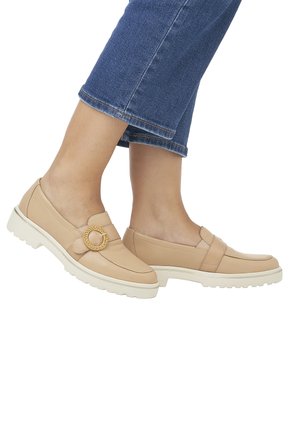 Des mocassins en cuir beige présentent une bande texturée et un accent circulaire, avec une semelle de traction blanche contrastante et un talon plat. Portés avec un denim court.