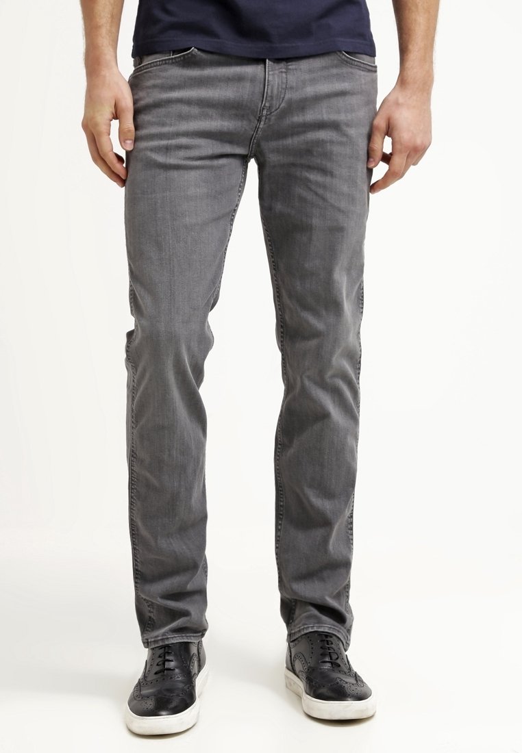 Homme portant un jean gris et des chaussures en cuir noires à lacets, debout sur un fond blanc, mains détendues le long du corps.