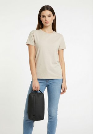 Joven con camiseta beige y vaqueros azules sosteniendo un neceser negro rectangular por el asa.