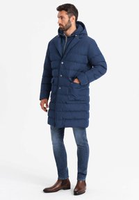 Abrigo de invierno acolchado en color navy con capucha, botones y bolsillos frontales. El abrigo presenta una textura acolchada y llega hasta la rodilla. Se combina con unos jeans y botas de tobillo.