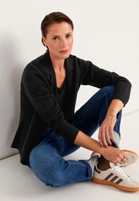 Sort tekstureret blazer over en sort top, parret med bl�å jeans rullet op ved håndleddene og beige sneakers med brune striber, siddende på gulvet.