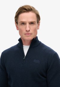 Maglione a zip colore blu navy realizzato in tessuto testurizzato, con colletto a costine e un logo discreto sul petto.