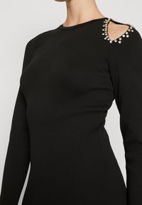Svart långärmad klänning med en figursydd silhuett, med en skuren axel prydd med metalliska nitar och en rund halsringning.