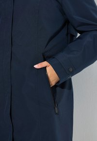 Mano con tono de piel claro en el bolsillo con cremallera de una chaqueta azul oscuro con puño abotonado.