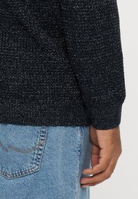 Tamsiai mėlynas megztinis su tekstūriniu raštu, su ribuotu rankogaliu ir apačia. Derinamas su šviesiai mėlynais džinsais.