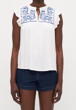 Biała bluzka z niebieskim haftowanym kwiatowym wzorem na dekolcie. Rękawy z falbanami. Połączona z ciemnoniebieskimi szortami dżinsowymi.