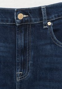 Jeans de ganga azul escuro com fecho de correr, botão metálico e costura branca. Apresentam um design com cinco bolsos, incluindo um bolso pequeno para moedas.