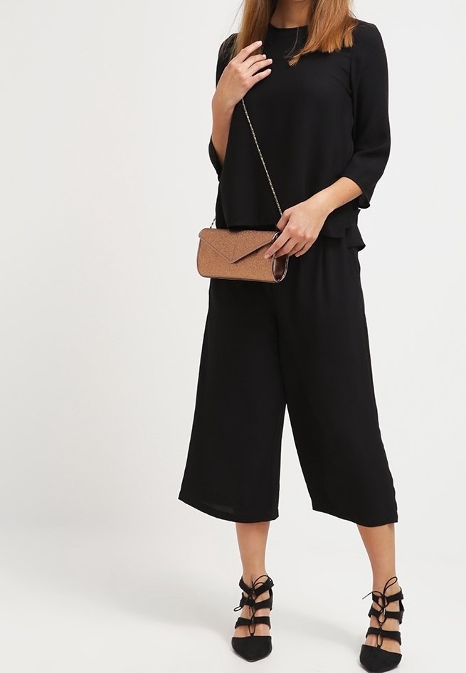 Czarny krótki kombinezon z lejącym się topem, zestawiony z czarnymi sandałami na cienkich paskach. Trzyma fakturowaną metaliczną kopertówkę.