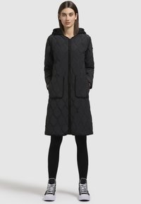 Cappotto lungo nero trapuntato con cappuccio, mentre presenta un motivo a rombi, chiusura frontale con zip e due tasche grandi. Indossato sopra leggins neri e sneakers.