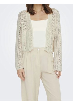 Femme portant un cardigan tricoté ouvert crème par-dessus un haut blanc à encolure carrée et un pantalon beige ample taille haute sur un fond uni.