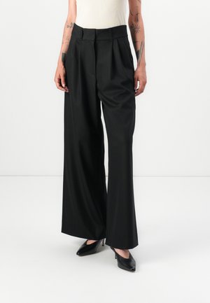 Pantalons larges plissés noirs à taille haute, fabriqués en tissu doux. Associés à des talons aiguilles noirs.