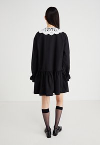 Rochie neagră cu mâneci lungi, volane plisate la poale și guler din dantelă albă. Asortată cu șosete până la genunchi și pantofi negri.