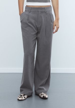 Personne portant un pantalon gris taille haute à jambes larges avec des plis, mains dans les poches, associé à des chaussures slip-on blanches et noires à motif.