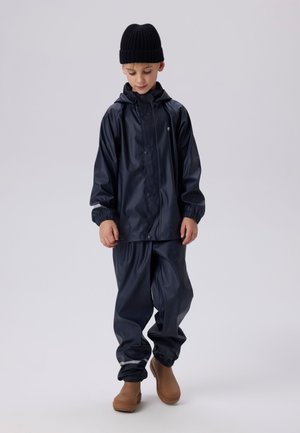 Marineblauwe waterdichte regenjas met capuchon en elastische manchetten, gecombineerd met bijpassende broek en beige waterdichte laarzen.