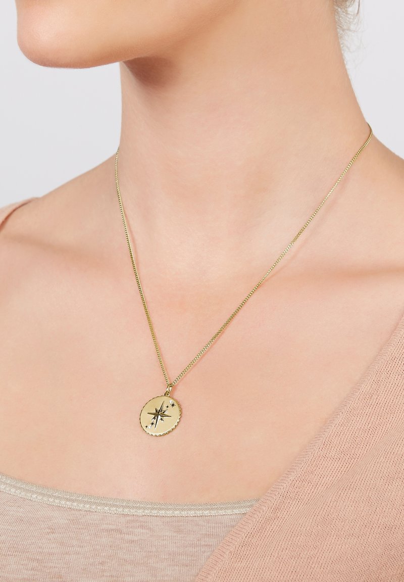 Collier en or avec un pendentif boussole rond porté autour du cou d'une femme, par-dessus un haut beige et un pull rose clair.