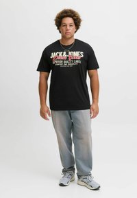 Schwarzes T-Shirt mit kurzen Ärmeln und einem Schriftzug in Weiß und Rot; kombiniert mit hellgrauen Jeans und grauen Sneakers. Lässige Passform und komfortable Materialien.