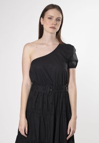 Vestido negro de un solo hombro con manga abullonada, que cuenta con un corsé ajustado y una falda de volantes. Viene con un cinturón a juego y detalles de bordado floral.