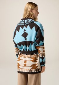 Cardigan lavorato a maglia con motivi geometrici blu, marroni e neri. Include un colletto con frange e una cintura in vita per un look aderente.