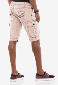 Tan Cargo-Shorts mit kontrastierenden dunklen Nähten, mehreren Seitentaschen und sichtbarem Markenlogo auf der Gesäßtasche. Getragen mit burgunderfarbenen Sneakern.